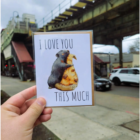 "I Love You This Much" Pizza Rat Card. A big rat hugging a giant slice of pizza. the maker is holding the card outside the subway.