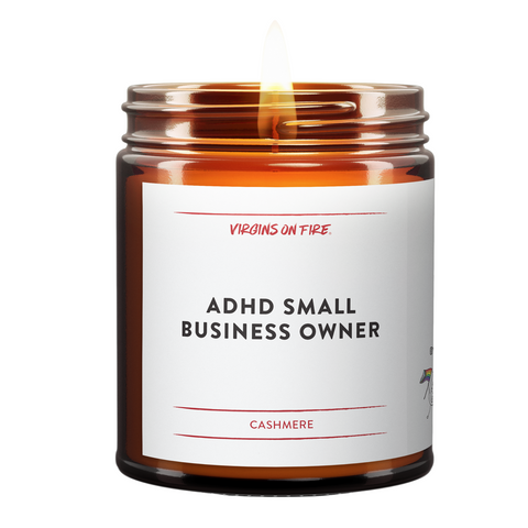 a burning candle in a brown amber jar. The name of the candle in black bold font is ADHD Small Business Owner. The scent is Cashmere. Red Virgins On Fire Logo.