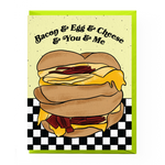BACON & EGG & CHEESE & YOU & ME GREETING CARD featuring a cartoon photo of a bacon egg and cheese sandwich. yellow background. white and black checker board foreground.