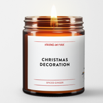 A holiday christmas candle called Christmas Decoration. it is Spiced Ginger. This is a photo of a lit candle. Big black font with the name. Virgins On Fire and Spiced Ginger in red.