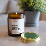 Candle jar with a humorous label on a table with a plant in the background. The label is Science Doesn't Care If You Believe In It.