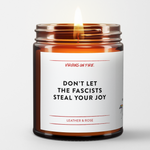 Burning candle. glass amber jar. white wraparound label with the text "Don't Let the Fascists Steal Your Joy."