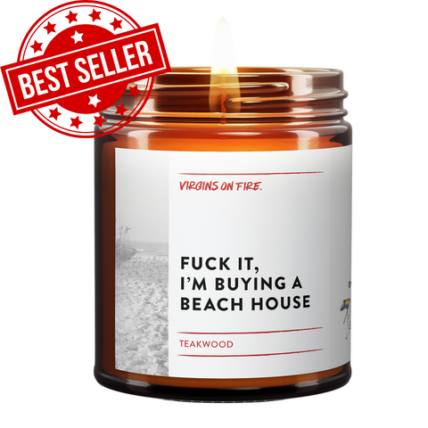 Amber glass candle jar with white label featuring black and white beach photo. Label reads 'Teakwood' scent by Virgins on Fire company. Contains mature language expressing desire to purchase a beach house. Small pride flag icon visible in corner.