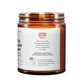 Back label of amber glass candle jar showing product details. 100% soy wax, handmade in Brooklyn, 45-50 hour burn time, non-toxic, 9oz size. Features Virgins on Fire Candle Co. branding, contact information, and cartoon mascot with rainbow flag. Includes note about being handmade by a gay man rather than a corporation."