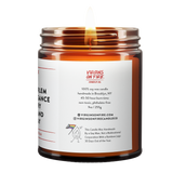 Back label of amber glass candle jar showing product details. 100% soy wax, handmade in Brooklyn, 45-50 hour burn time, non-toxic, 9oz size. Features Virgins on Fire Candle Co. branding, contact information, and cartoon mascot with rainbow flag. Includes note about being handmade by a gay man rather than a corporation."