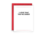 red envelope, white background greeting card with black text that reads, "I love that you're sober" in bold lettering. this is a message of sobriety support from virgins on fire candle co - a sober run business.