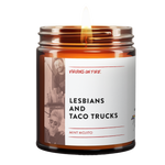 Amber glass candle from Virgins On Fire Candle Co., a funny gay-owned Brooklyn, NY candle business. Label reads 'Lesbians and Taco Trucks' with mint mojito scent. Features black and white lifestyle photographs and lit wick. Small pride flag icon visible.