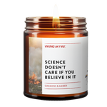 Lit amber glass candle with white label featuring 'Science Doesn't Care If You Believe In It' text and background image of flames, scented with oakmoss and amber by Brooklyn-based Virgins on Fire