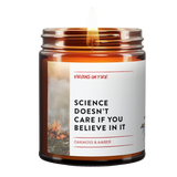 Lit amber glass candle with white label featuring 'Science Doesn't Care If You Believe In It' text and background image of flames, scented with oakmoss and amber by Brooklyn-based Virgins on Fire
