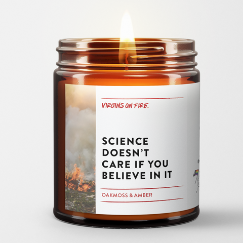 Lit amber glass candle with white label featuring 'Science Doesn't Care If You Believe In It' text and background image of flames, scented with oakmoss and amber by Brooklyn-based Virgins on Fire