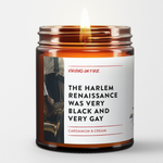 Amber glass candle jar with white label featuring vintage jazz musician photo. Label reads 'The Harlem Renaissance Was Very Black and Very Gay' with Cardamom & Cream scent description. Small pride flag icon visible. Made by Virgins on Fire company.