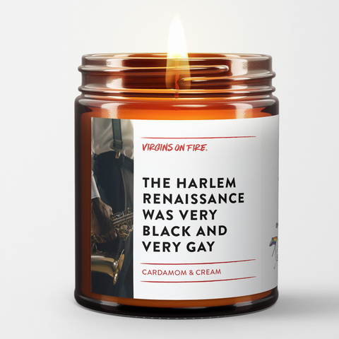 Amber glass candle jar with white label featuring vintage jazz musician photo. Label reads 'The Harlem Renaissance Was Very Black and Very Gay' with Cardamom & Cream scent description. Small pride flag icon visible. Made by Virgins on Fire company.