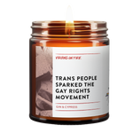 a lit amber jar candle called, "Trans people sparked the gay rights movement". Photo of a brick on the side. Gin and cypress scented.