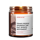 a lit amber jar candle called, "Trans people sparked the gay rights movement". Photo of a brick on the side. Gin and cypress scented.