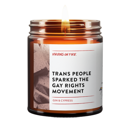 a lit amber jar candle called, "Trans people sparked the gay rights movement". Photo of a brick on the side. Gin and cypress scented.