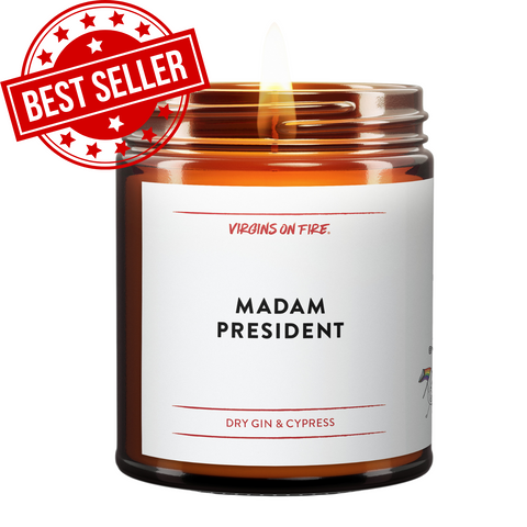 An amber brown jar with a lit candle. The text is "MADAM PRESIDENT" it is Gin & Cypress scented Soy Wax. This candle is for the 2024 Election. Has Best Seller in the corner.