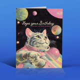 a cartoon cat - pink and gray - in a space suit floating in space around pastel colored planets. It says in script, "hope your birthday is out of this world."