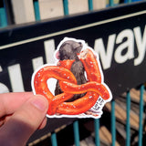 the maker of the sticker holding the sticker. It is of a gray rat eating a pretzel. cute cartoon style new york city subway sticker.