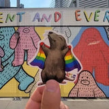 the maker of the sticker holding his sticker. cute cartoon rat holding a rainbow pride flag. Against a colorful urban background.
