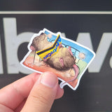 cute cartoon style nyc subway rat laying on a towel that says Coney Island and using an MTA subway card to direct sunlight onto themselves like a reflector. funny and cute. The maker is holding it for the photo.