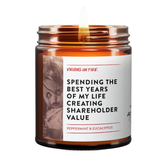 A lit amber glass jar candle with a white label reading 'SPENDING THE BEST YEARS OF MY LIFE CREATING SHAREHOLDER VALUE' and 'PEPPERMINT & EUCALYPTUS' by Virgins On Fire, shown on a white background. there is also an illustration on the left of the label of a wounded doll
