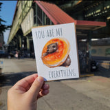 a greeting card outside of a subway station. it says You are my everything. there is a rat stuck in a bagel.