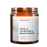 Amber glass candle jar with white label featuring black and white beach photo. Label reads 'Teakwood' scent by Virgins on Fire company. Contains mature language expressing desire to purchase a beach house. Small pride flag icon visible in corner.