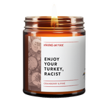 Enjoy Your Turkey Racist is the name of a candle from Virgins On Fire Candle Co. This brown amber jar candle is lit. There is a photo of a thanksgiving dinner on the label. it says, "Enjoy Your Turkey, Racist." It is a cranberry and pine scent. 