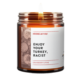 Enjoy Your Turkey Racist is the name of a candle from Virgins On Fire Candle Co. This brown amber jar candle is lit. There is a photo of a thanksgiving dinner on the label. it says, "Enjoy Your Turkey, Racist." It is a cranberry and pine scent. 