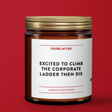 Gold Capped soy wax candle in brown amber jar with the text, "Excited to Climb the Corporate Ladder then die." Amber and Driftwood scent. From Virgins On Fire Candle Co., a gay-owned small business in Brooklyn, NY. Red background.