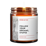 a brown amber jar. lit candle. text reads, "Follow your dreams, stupid." Smoked Oud scented. Includes a photo of an ambitious girl holding a wooden airplane. 