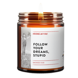 a brown amber jar. lit candle. text reads, "Follow your dreams, stupid." Smoked Oud scented. Includes a photo of an ambitious girl holding a wooden airplane. 