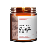 Foxy ladies doin a lil afternoon shoppin is the name of a candle from virgins on fire candle co. This is a brown amber jar candle. lit. has that phrase. woman smelling rosemary in photo. rosemary and sage scented.