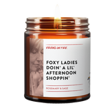 Foxy ladies doin a lil afternoon shoppin is the name of a candle from virgins on fire candle co. This is a brown amber jar candle. lit. has that phrase. woman smelling rosemary in photo. rosemary and sage scented.