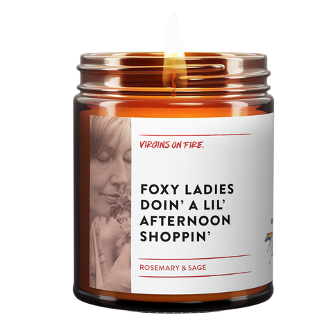 Foxy ladies doin a lil afternoon shoppin is the name of a candle from virgins on fire candle co. This is a brown amber jar candle. lit. has that phrase. woman smelling rosemary in photo. rosemary and sage scented.