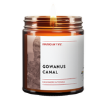 Gowanus Canal scented candle by queer-owned gay Brooklyn candlemaker Virgins On Fire. Features Cashmere & Tonka fragrance in 9oz amber glass jar. Funny Minimalist white label design with red accents. Lit wick showing warm flame glow.